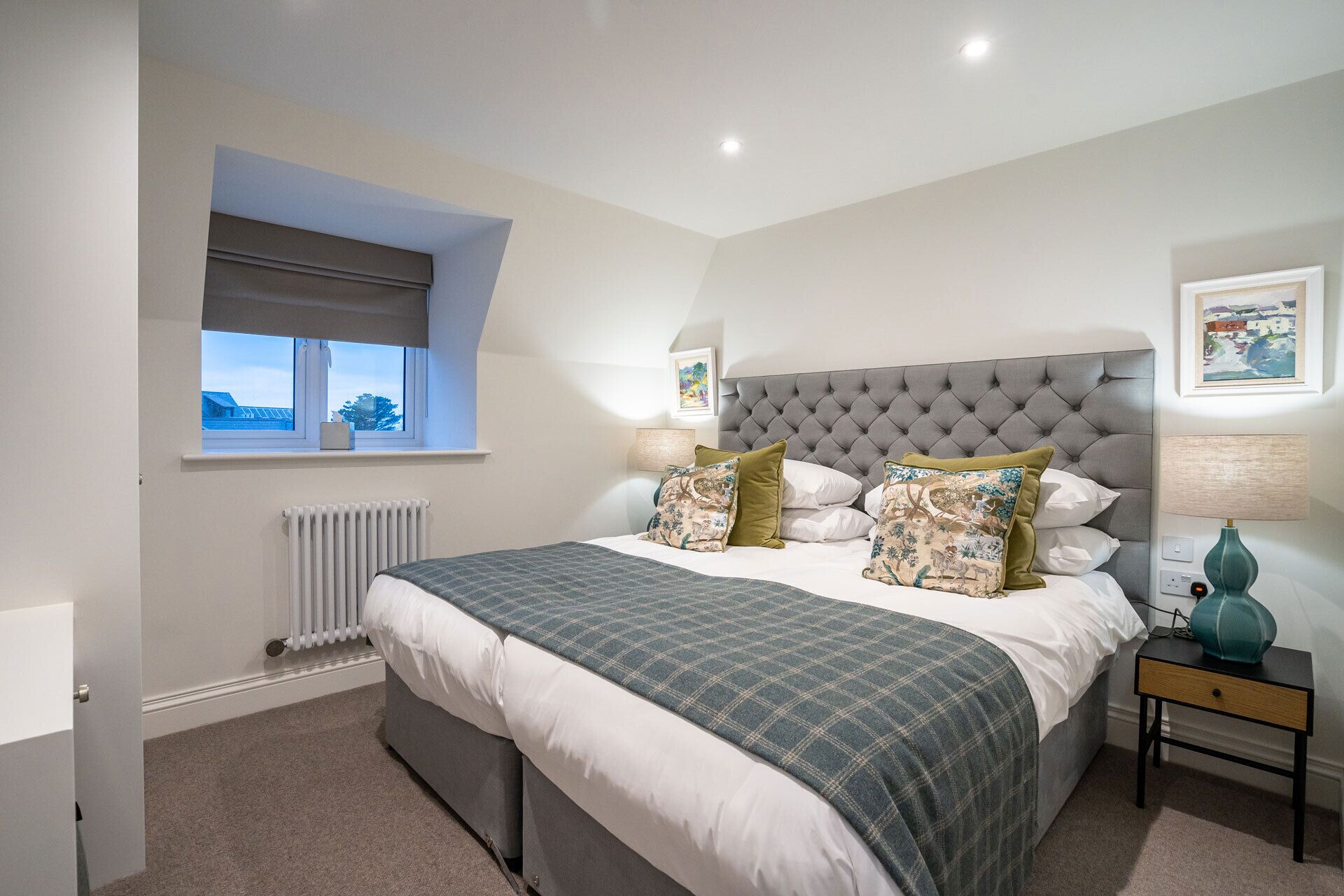 A bedroom with a large bed, a window with a view, a radiator, and a bedside table with a lamp.