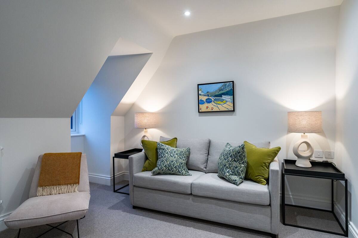 A modern living room with a grey sofa, patterned cushions, and two side tables with lamps. A painting of a beach scene hangs on the wall.