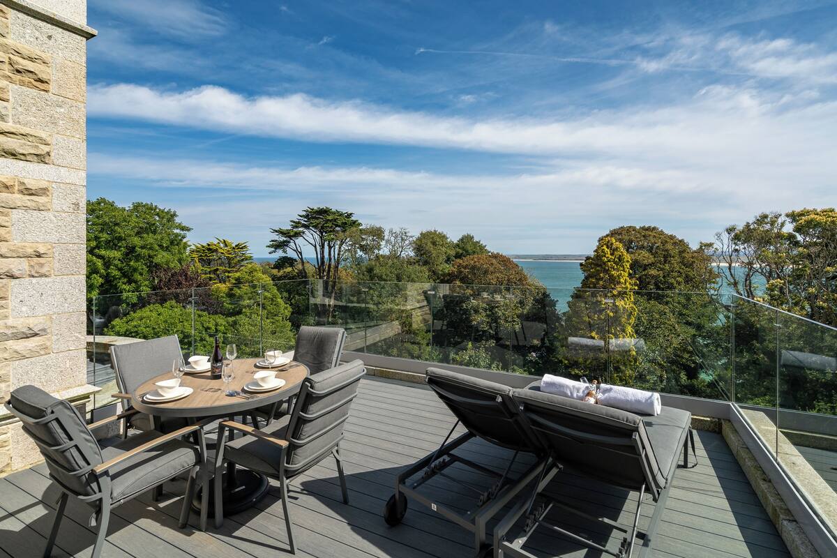 A balcony with a table set for dining, lounge chairs, and a view of the sea.