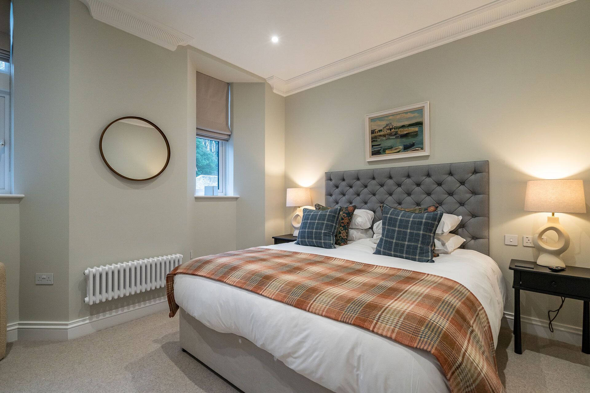 A bedroom with a large bed, two bedside tables, a round mirror, a window, and a painting on the wall.