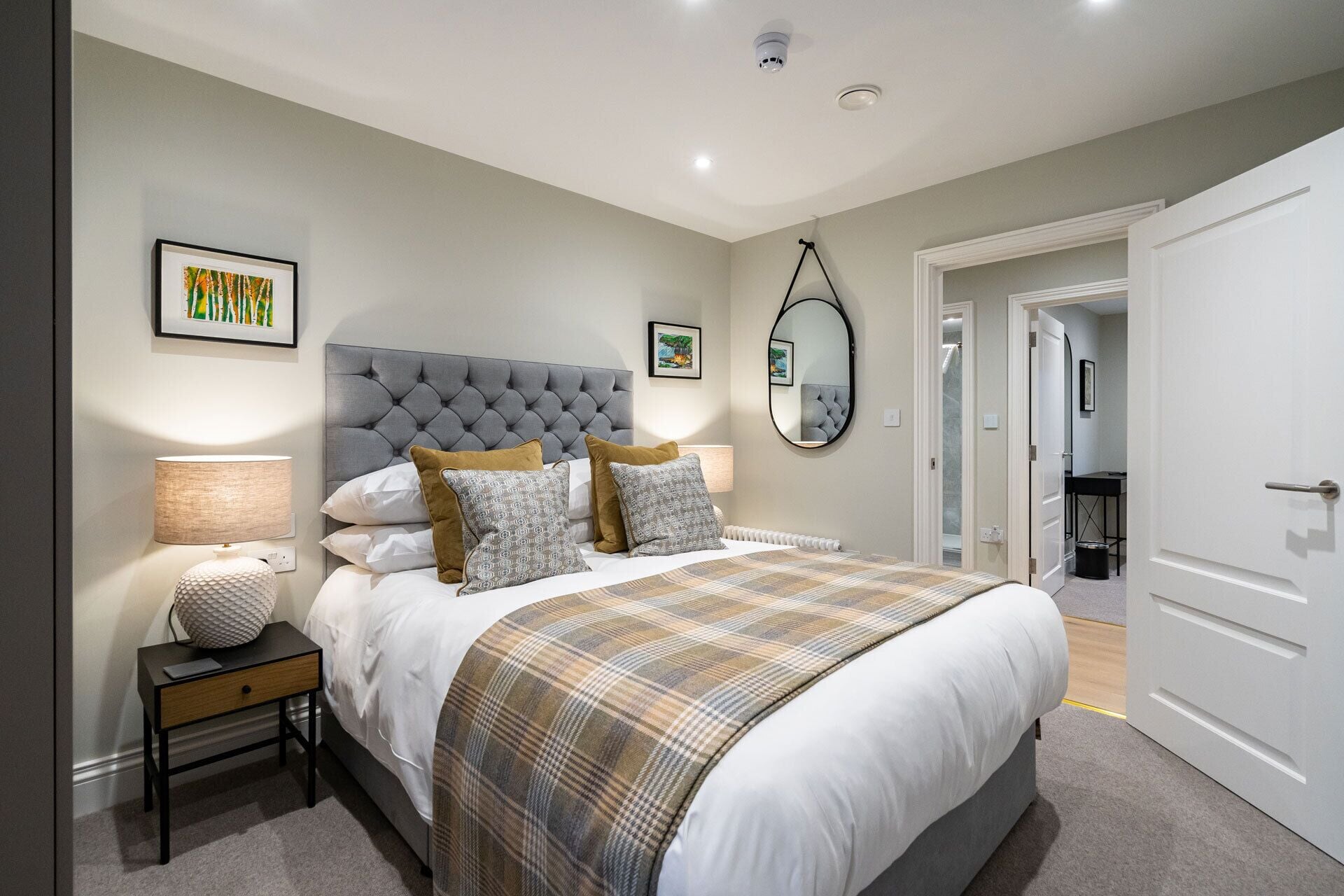 A bedroom with a large bed, a bedside table with a lamp, a mirror, and artwork on the walls.