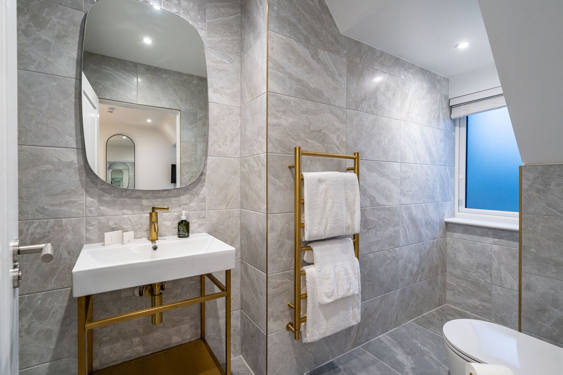 A modern bathroom with a white sink, gold fixtures, a towel rack, and a window with a view.