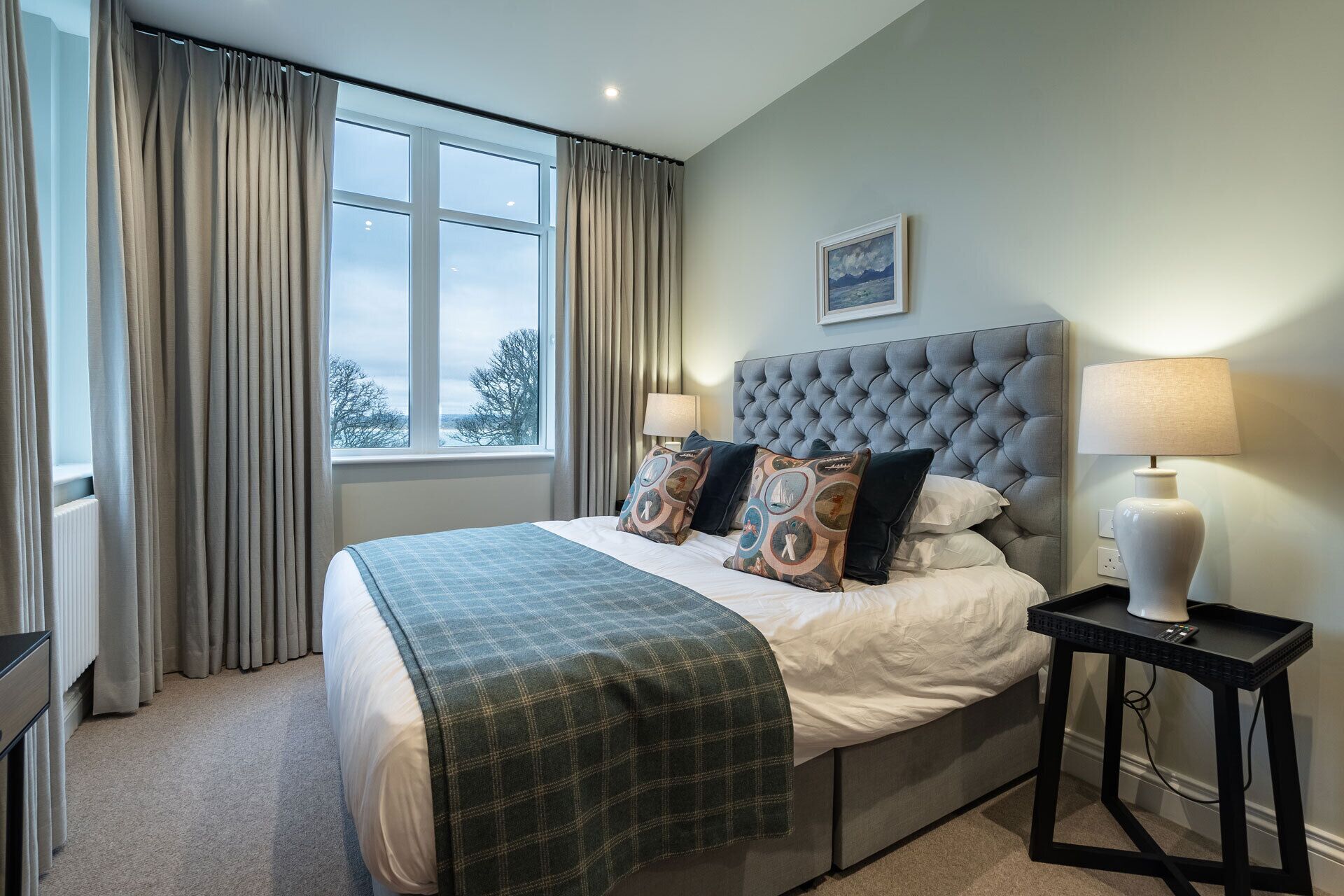 A bedroom with a large bed, two bedside lamps, a window with curtains, and a framed picture on the wall.