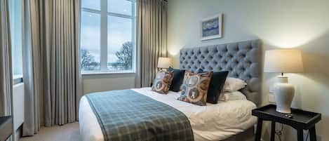 A bedroom with a large bed, two bedside lamps, a window with curtains, and a framed picture on the wall.