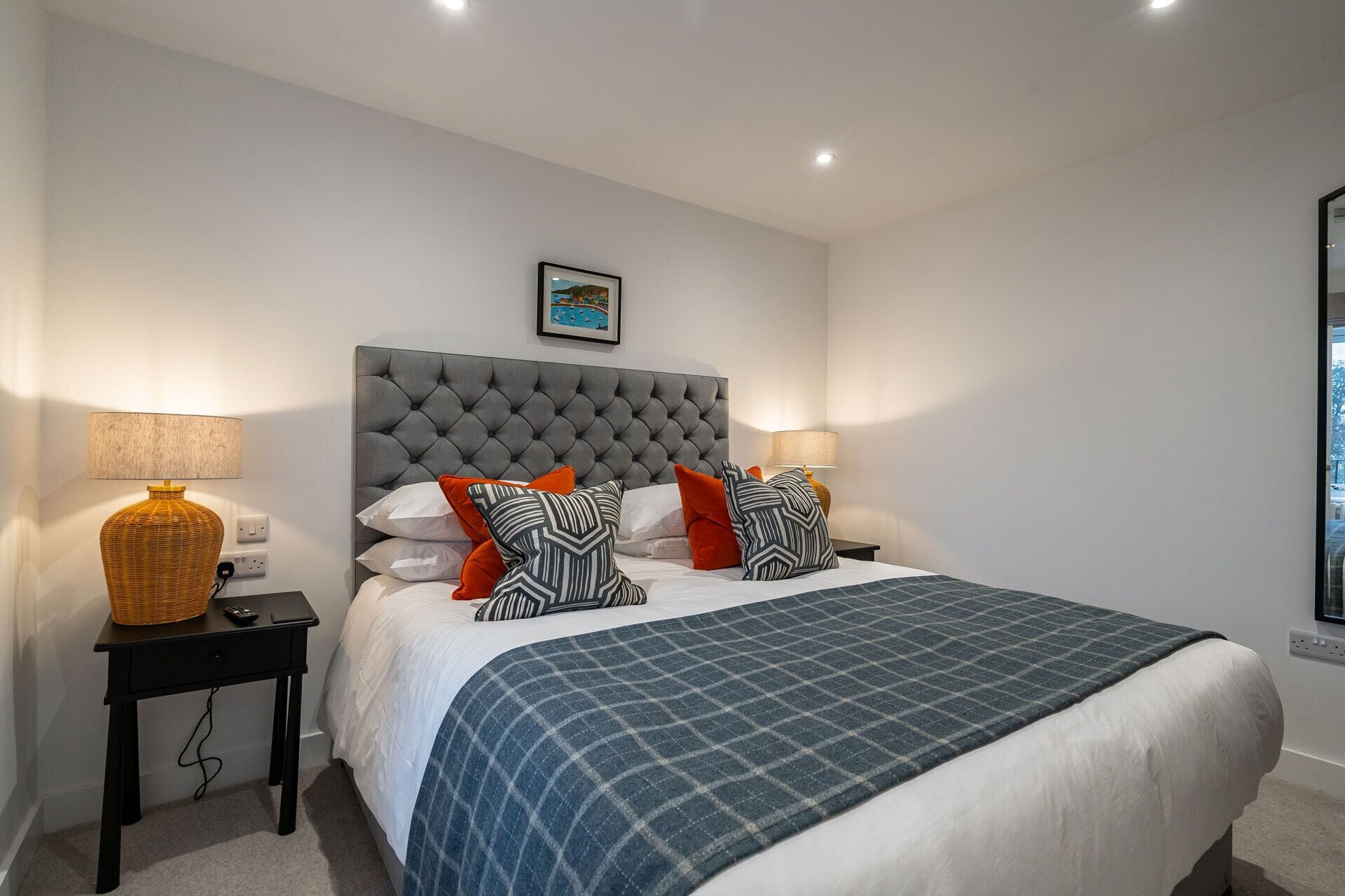 A neatly made bed with a grey tufted headboard, white bedding, and a blue plaid throw. There are two bedside tables with lamps and a framed picture above the bed.