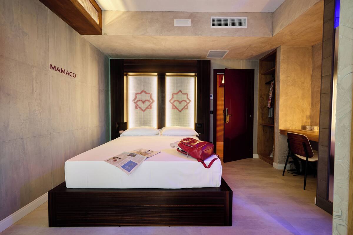 A hotel room with a bed, a wooden headboard, a desk with a chair, and a wardrobe.
