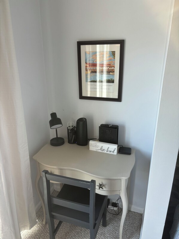 A small, white desk with a black chair, a framed picture, a lamp, a speaker, and a sign saying "In a world where you can be anything... be kind." - Charming studio in lovely Emerald Isle with beach access (Emerald Isle)