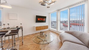 Un salon moderne avec une table basse en verre, un canapé, un téléviseur fixé au mur et une vue sur la ville grâce à de grandes fenêtres.