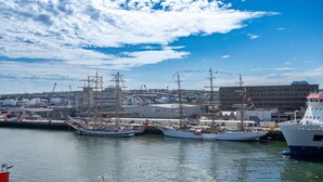 Un port où sont amarrés de nombreux voiliers, un ciel dégagé et, au loin, des bâtiments industriels.