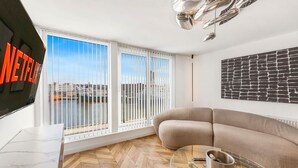 Un salon moderne avec un canapé, un téléviseur et une vue sur un plan d’eau, grâce à de grandes fenêtres.