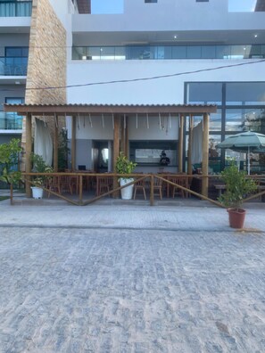 Un área de estar al aire libre con mesas y sillas de madera, plantas en macetas y un edificio moderno al fondo.