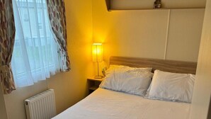 A hotel room with a bed, a nightstand with a lamp, and a window with curtains. - 3 Bed Caravan - Sleeps 6 - Pets - 5 min to Beach (Abergele)