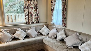 A corner sofa with multiple cushions, a wooden bench, and a window with patterned curtains. - 3 Bed Caravan - Sleeps 6 - Pets - 5 min to Beach (Abergele)