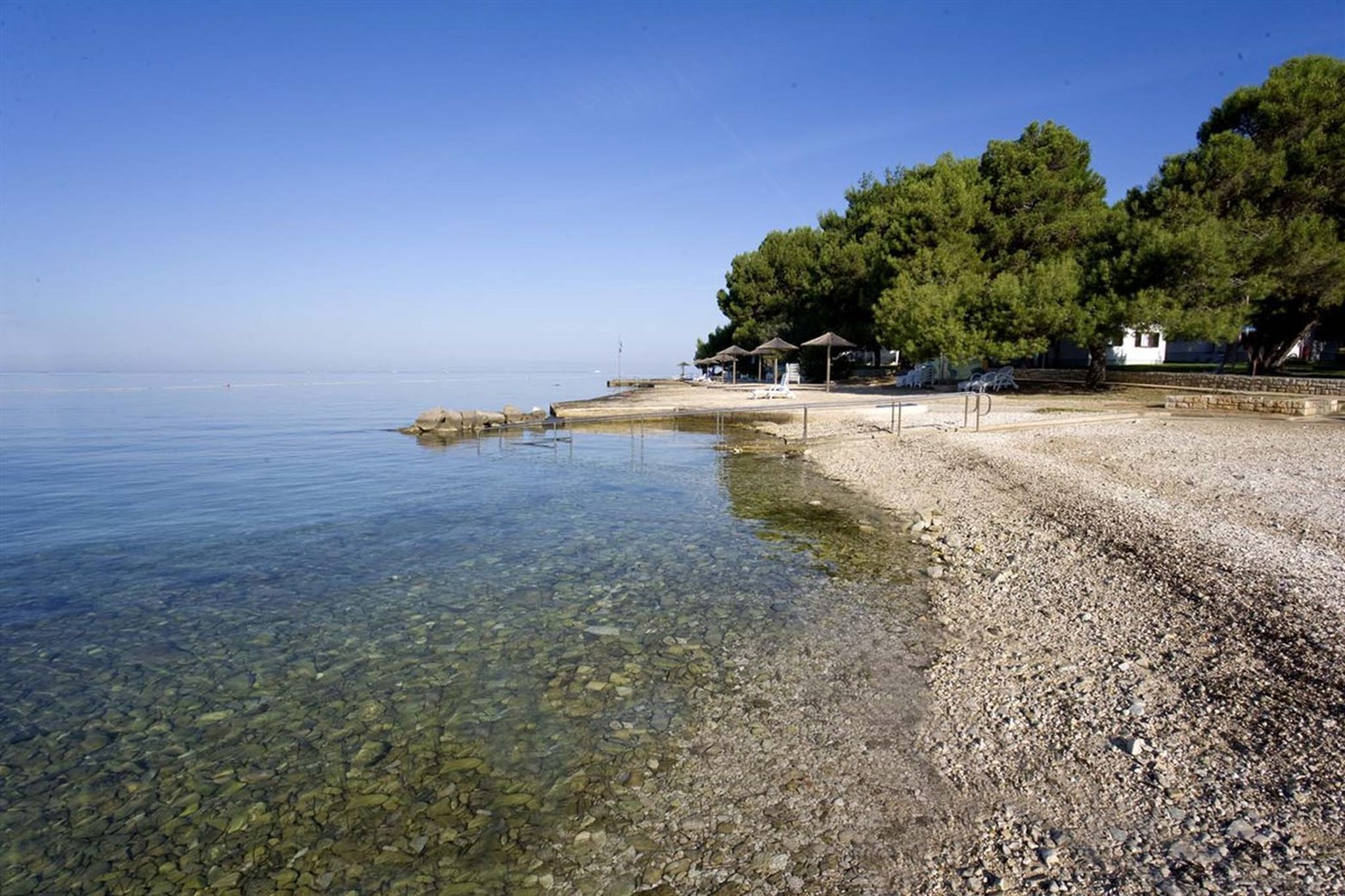 A pebble beach with clear water, a few umbrellas, and a building with a white roof.