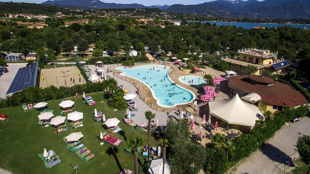 An aerial view of a resort with a large swimming pool, sun loungers, and a view of the surrounding mountains and sea. - Vacation resort Baia Verde Mobile Home Suite Deluxe for 5 persons (Manerba del Garda)