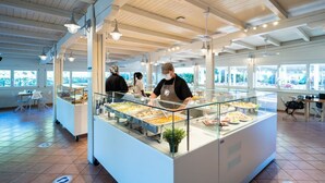 Un espace buffet comprenant plusieurs stands de nourriture, un membre du personnel assurant le service, et un espace repas avec des tables et des chaises.