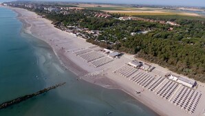 Une vue aérienne d’une plage avec des rangées de chaises longues et de parasols, une jetée qui s’avance dans l’eau, et un quartier résidentiel avec des maisons et de la végétation en arrière-plan.