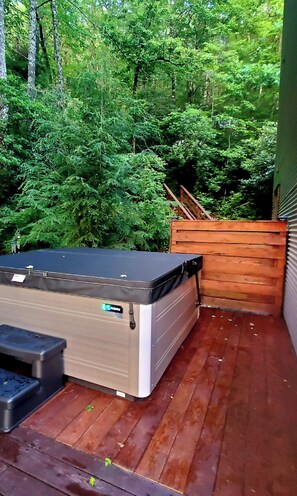A wooden deck with a mattress and a small table, surrounded by dense green foliage.