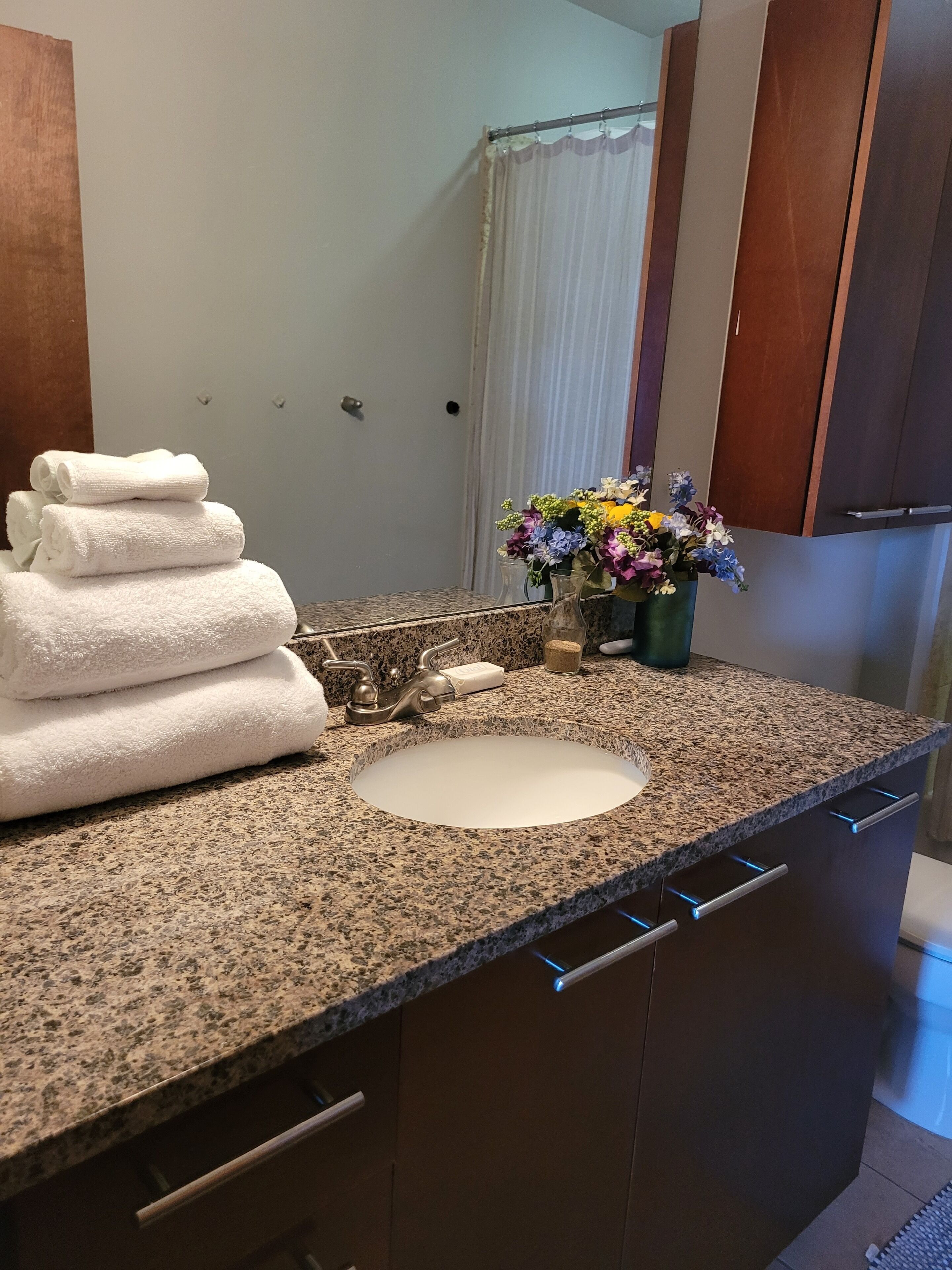 Ein Badezimmer mit Granitplatte, Waschbecken, Spiegel, gestapelten Handtüchern und einer Vase mit Blumen.