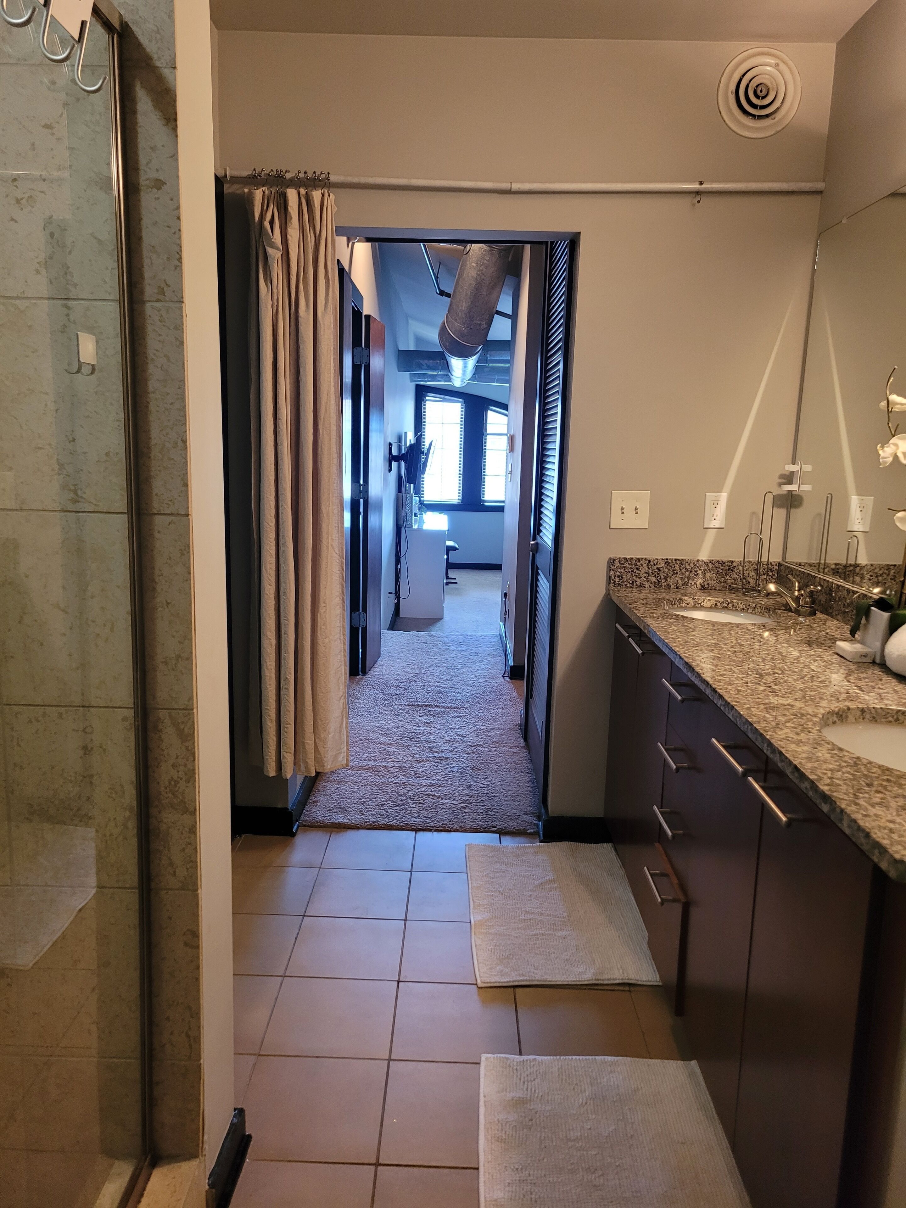Ein Badezimmer mit Dusche, Waschbecken und Spiegel. Ein Handtuchhalter ist vorhanden und ein Handtuch hängt daran. Der Boden ist mit Fliesen ausgelegt und es gibt ein Fenster mit Jalousien.