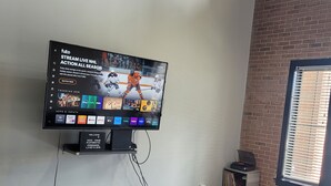A living room with a flat-screen TV mounted on the wall displaying a sports channel. There is a small table with a vase and a cushion, a window with blinds, and a brick wall.