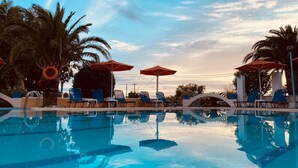 A swimming pool with clear blue water, surrounded by lounge chairs and umbrellas, palm trees, and a clear sky.