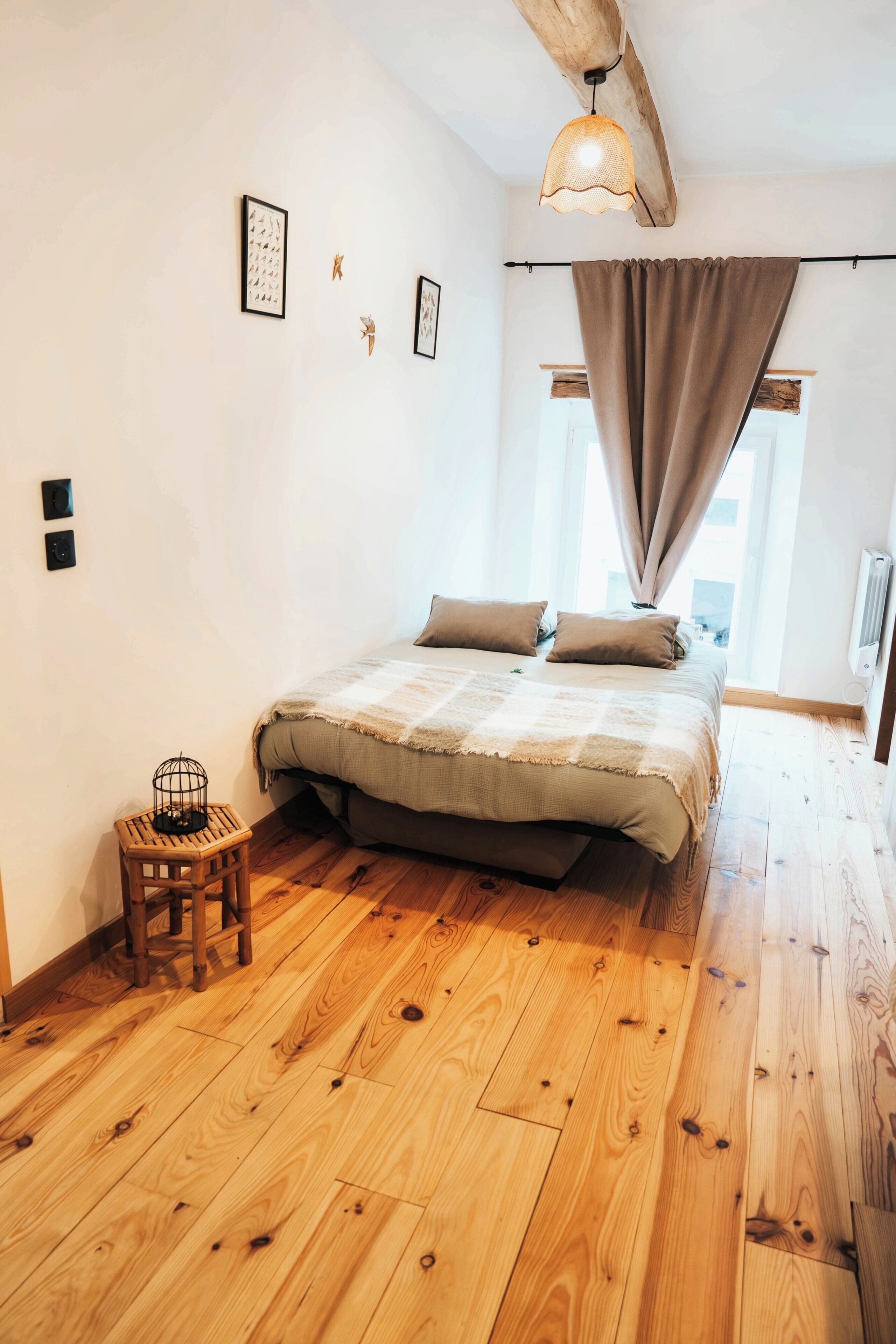 Een slaapkamer met een houten vloer, een bed met een beige deken, een klein houten krukje en een hanglamp.