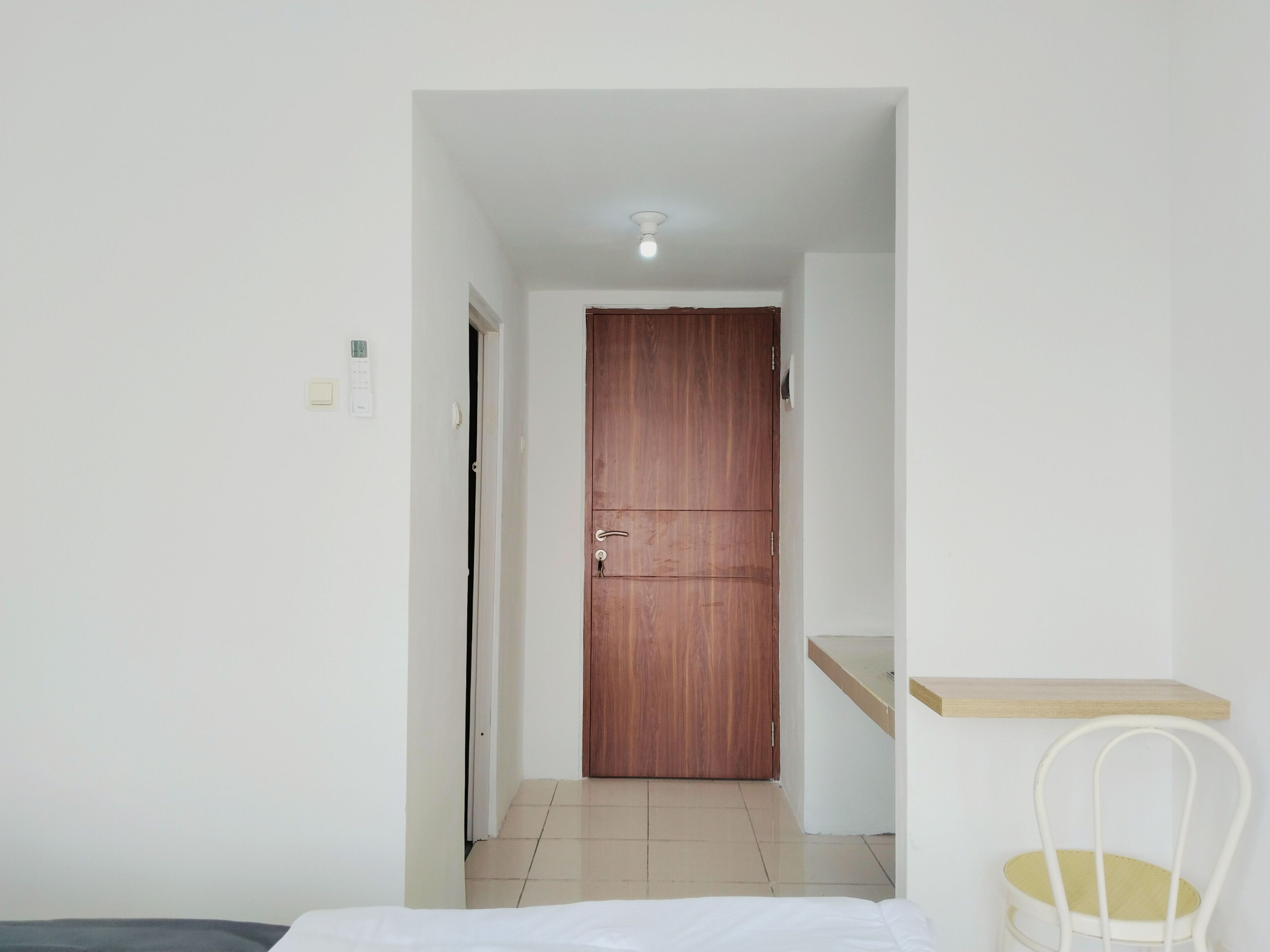 Habitación de hotel con puerta de madera, cama blanca, una pequeña silla blanca y un estante de madera.