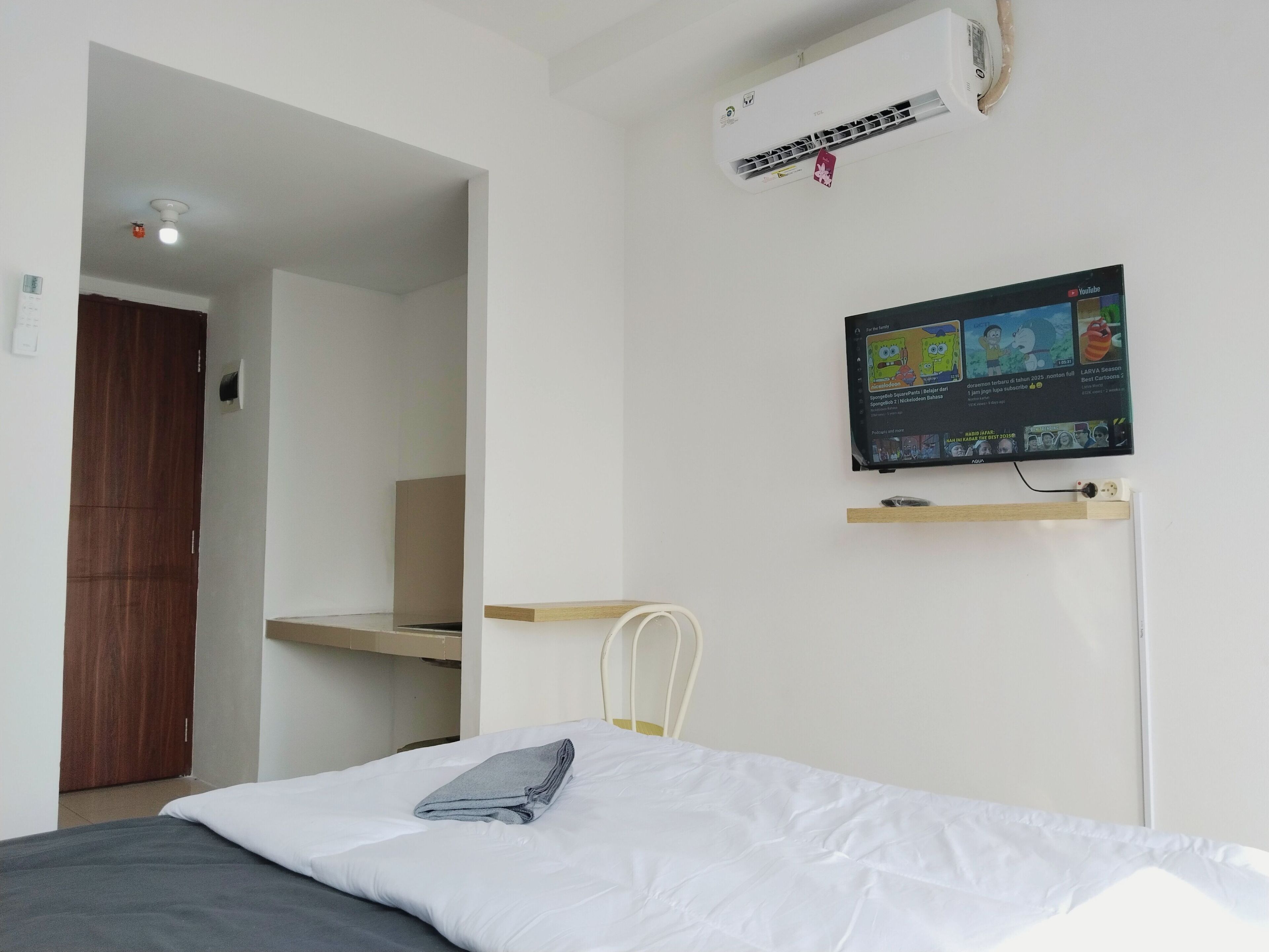 A bedroom with a bed, a TV mounted on the wall, a wooden door, and an air conditioning unit.
