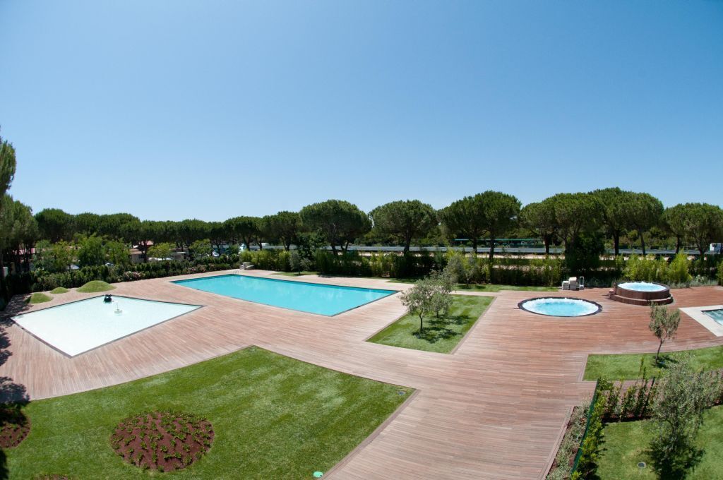 Ein Poolbereich mit rechteckigem Pool, Whirlpool und einem runden Whirlpool, umgeben von Holzdeck und Rasenflächen.