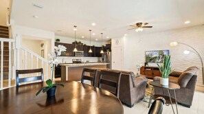 Una cocina moderna con armarios oscuros, una isla central, iluminación colgante y un comedor con una mesa y sillas marrones.