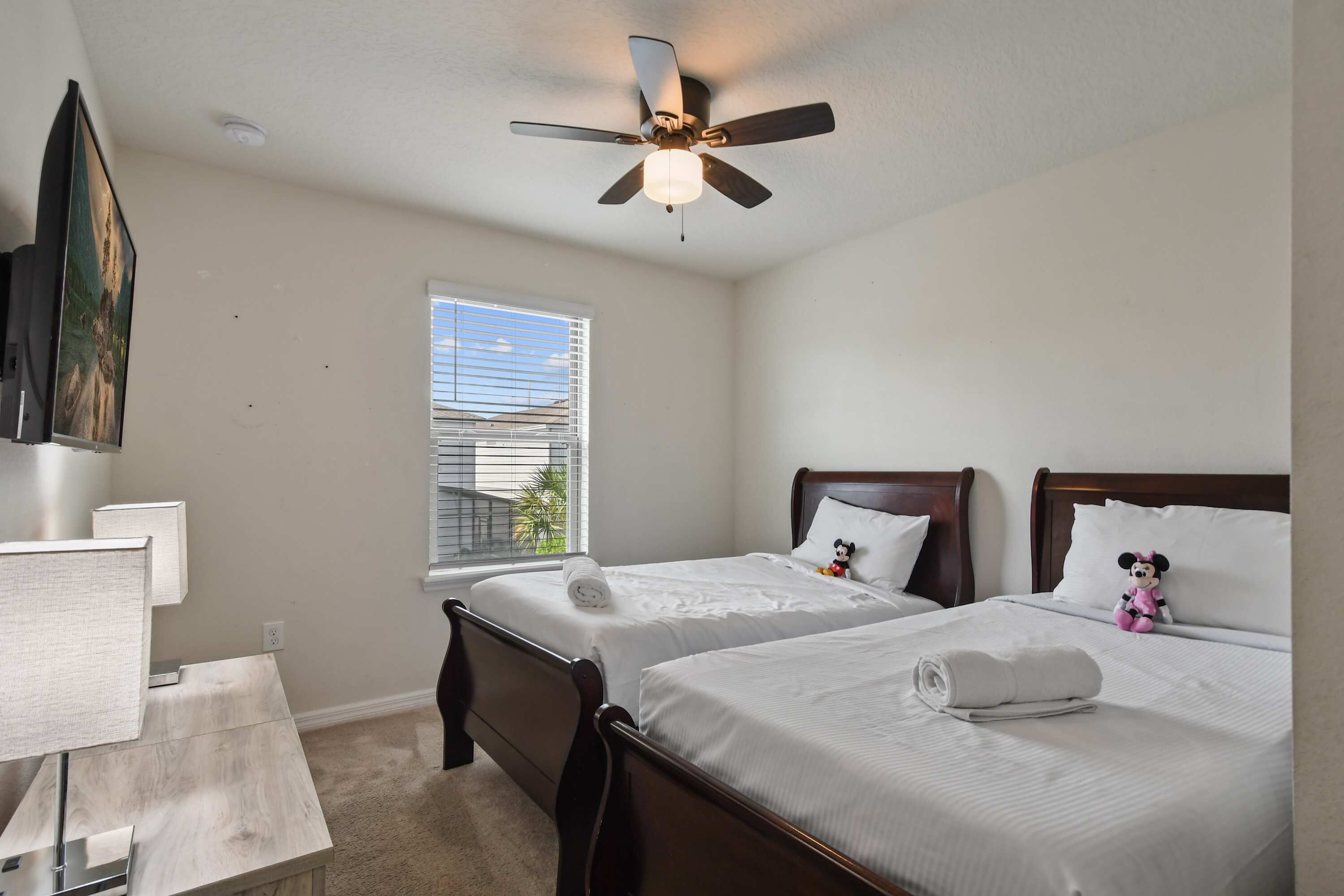 A hotel room with two beds, a ceiling fan, a window with blinds, a desk with a lamp, and a TV mounted on the wall.