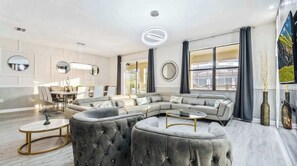Un salon moderne avec un canapé d’angle, des poufs ronds, un coin repas et de grandes fenêtres.