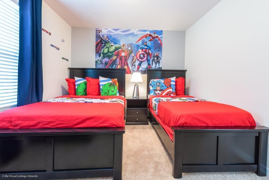 Ein Schlafzimmer mit zwei Betten, jeweils mit roter Bettwäsche und bunten Kissen. An der Wand befindet sich ein Wandbild mit Superhelden und ein Nachttisch mit einer Lampe.