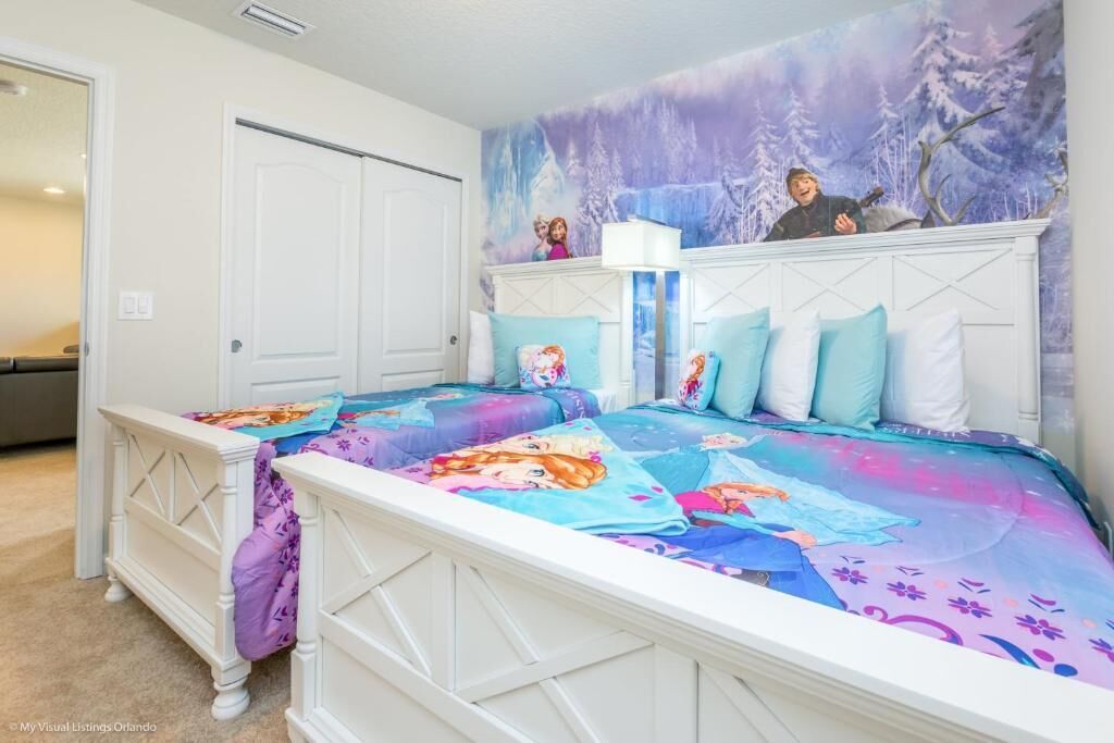 A bedroom with two beds, each with a colorful bedspread featuring cartoon characters. There is a white headboard with a decorative pattern, and a mural of a snowy landscape on the wall behind the beds.