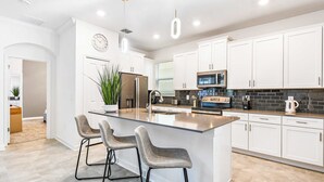 Cocina moderna con armarios blancos, una isla central con taburetes de bar, electrodomésticos de acero inoxidable y un salpicadero de azulejos negros.