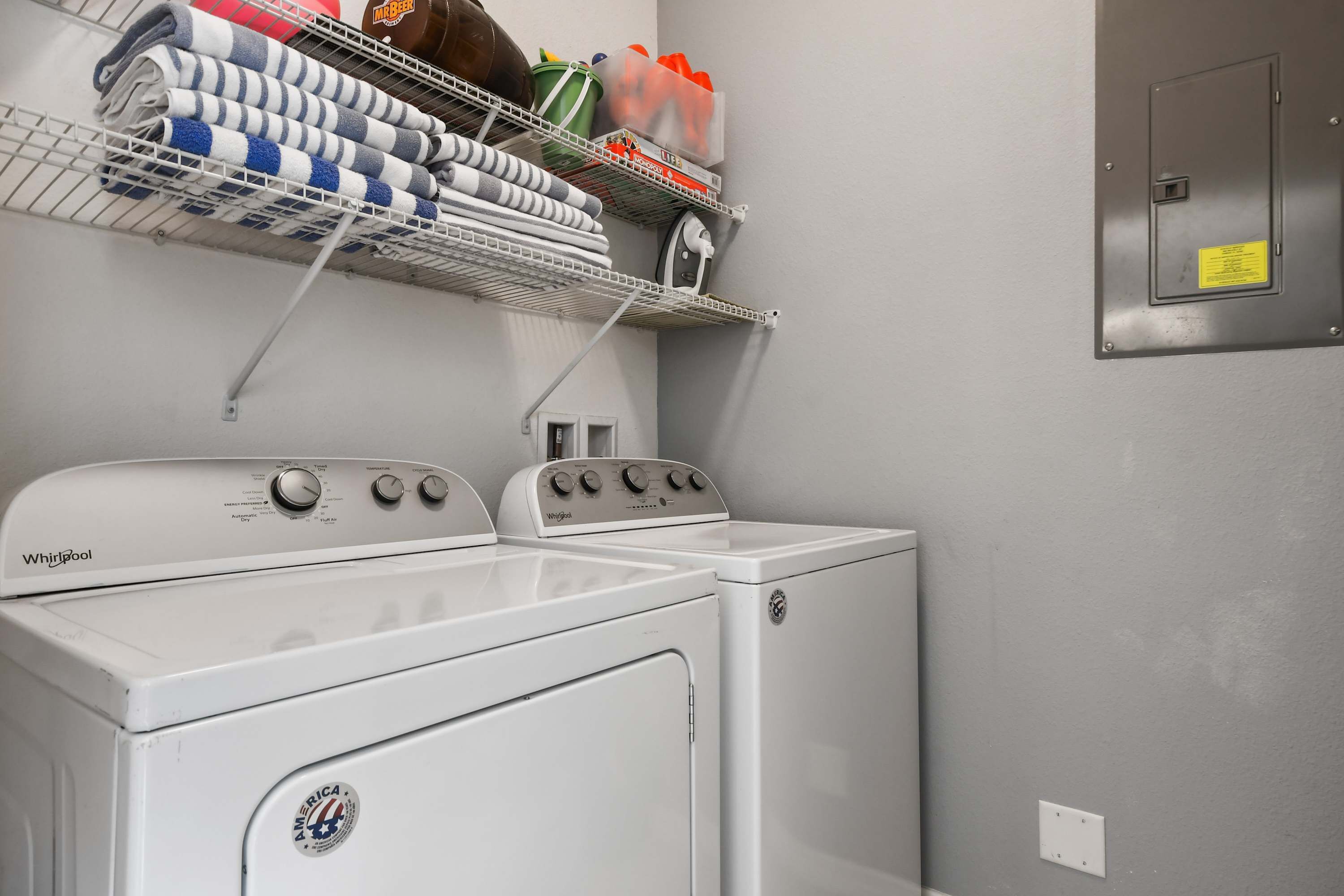 A laundry area with two washers and dryers, a metal shelf above holding folded towels and cleaning supplies, and a utility panel on the wall.