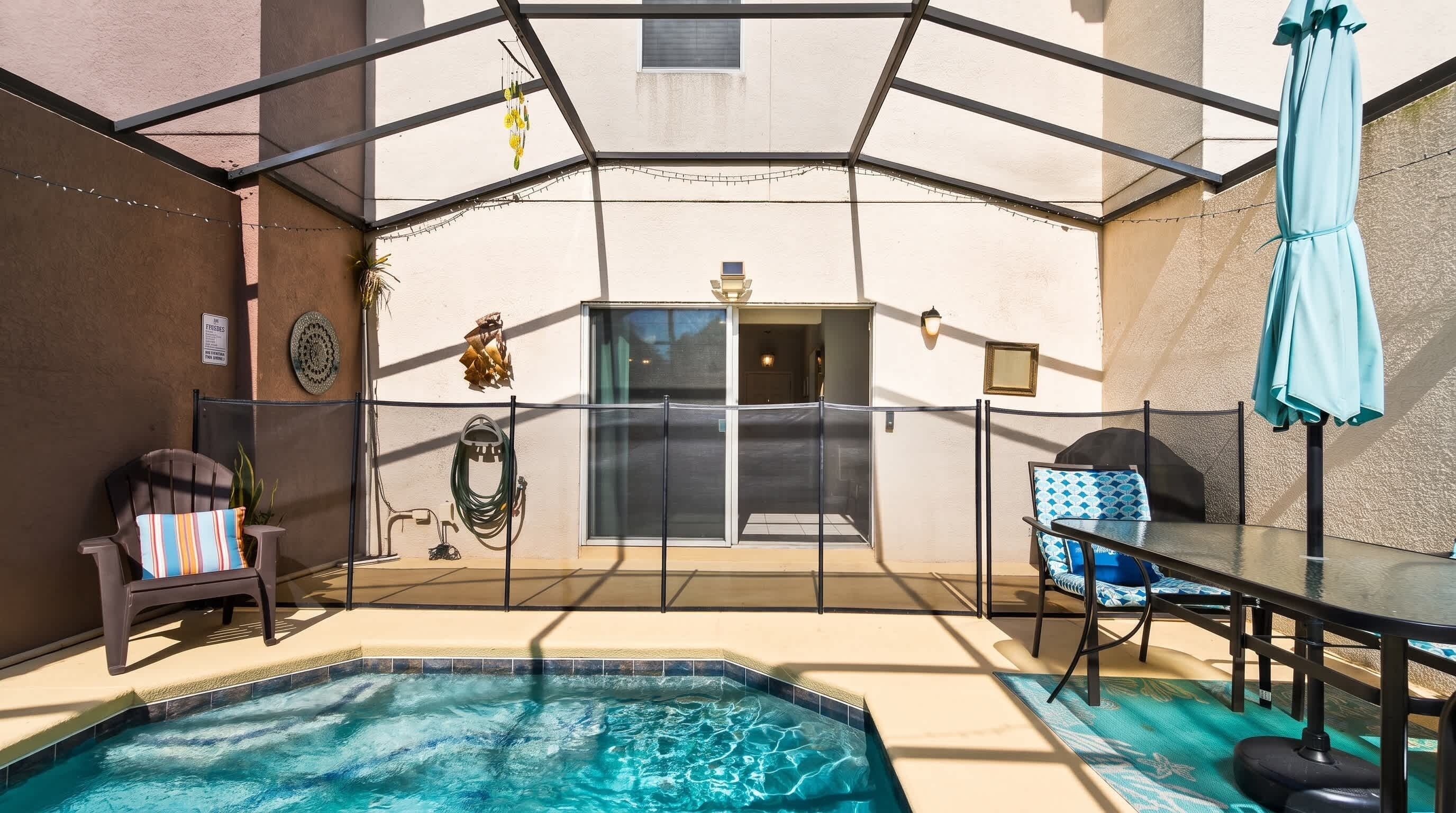 A pool area with a glass enclosure, a table with chairs, a chair with a striped cushion, and a blue umbrella.
