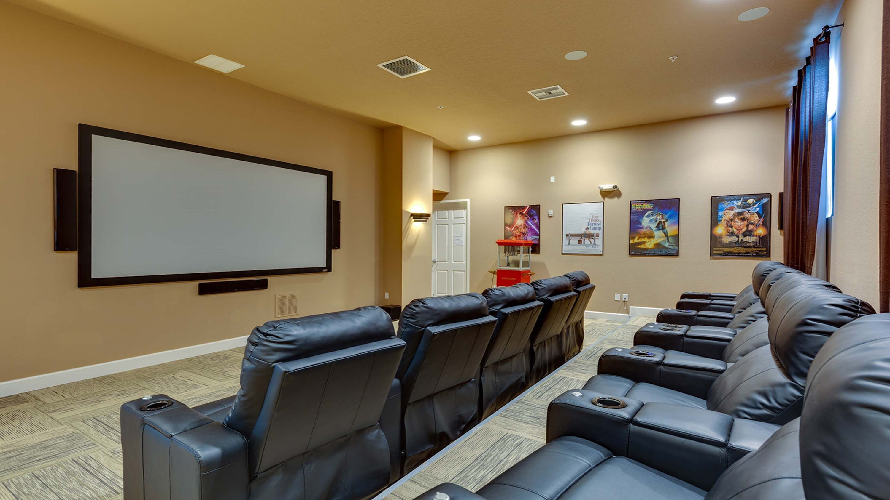 A home theater room with a large screen, multiple rows of leather recliner seats, and several movie posters on the wall.