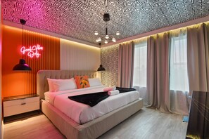 A modern bedroom with a geometric patterned ceiling, a neon sign, a large bed with pillows, and a bedside table.