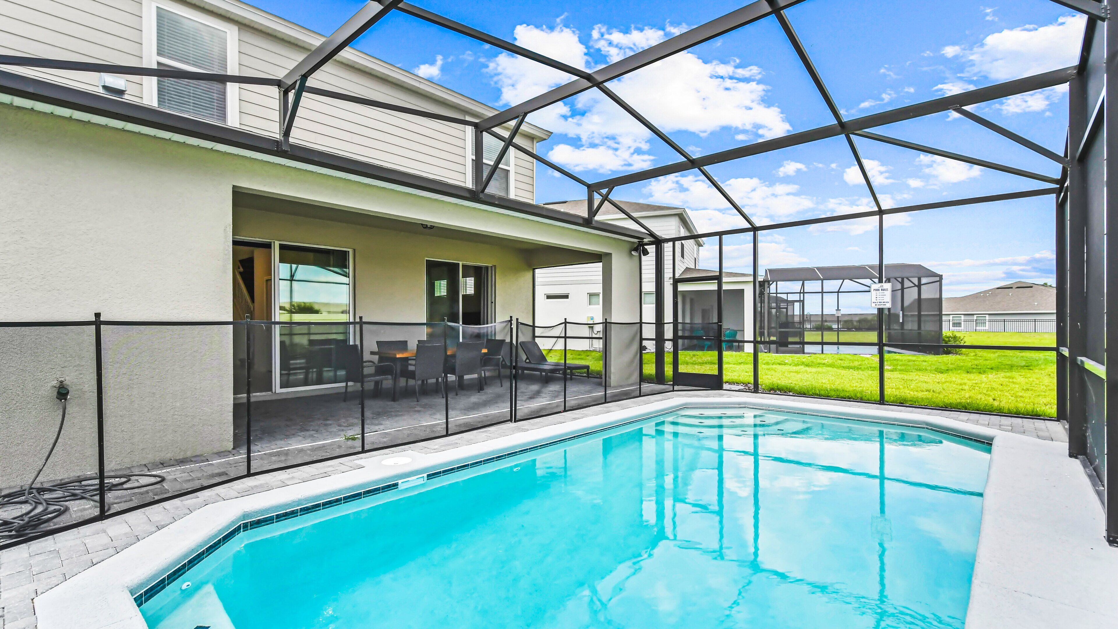 Ein Schwimmbecken mit Glasveranda, eine Terrasse mit Stühlen und Tisch sowie ein Blick auf eine Rasenfläche und Gebäude im Hintergrund.