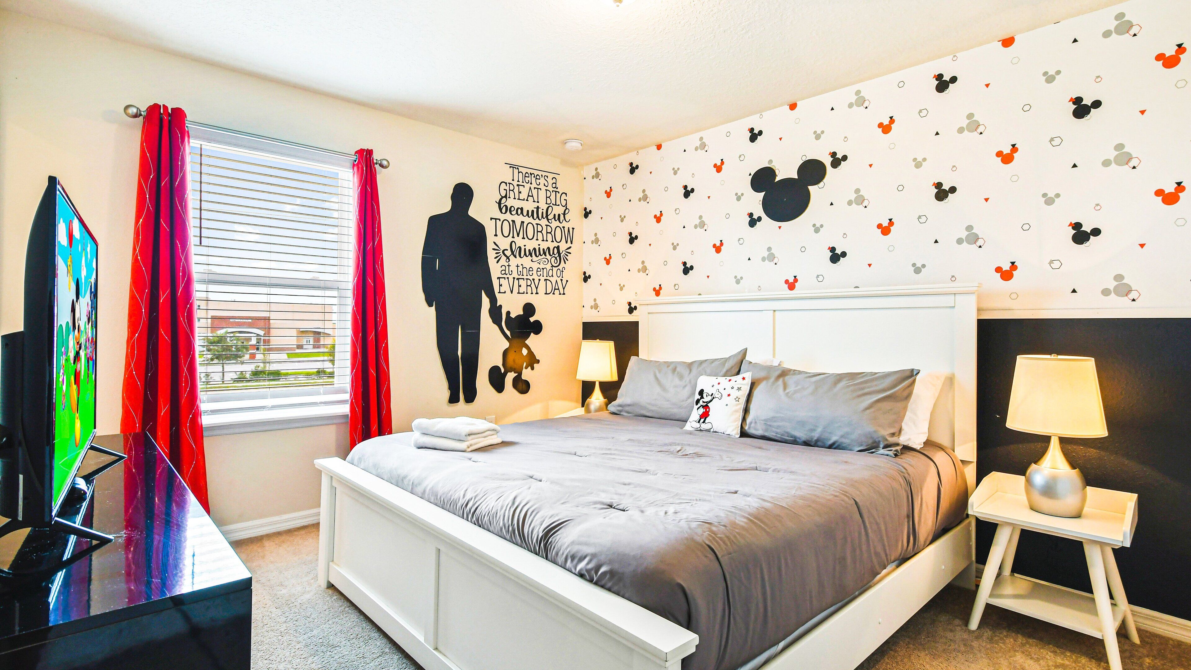 Une chambre avec un lit, une télévision, une fenêtre avec des rideaux, un mur décoré d’un autocollant de Mickey Mouse et une citation inspirante.