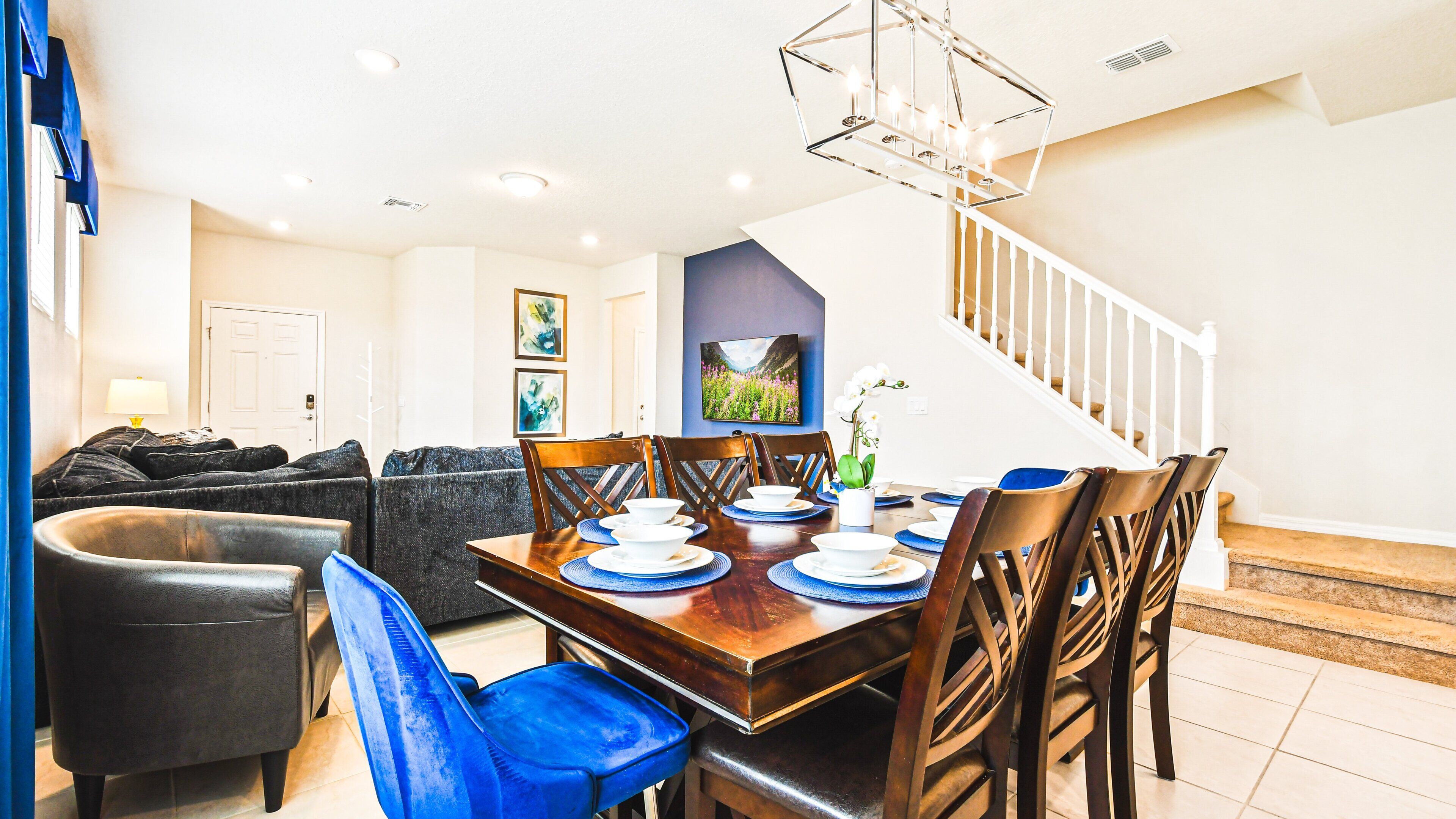 A dining area with a wooden table set for four, surrounded by blue chairs. A staircase is visible in the background leading to an upper floor.