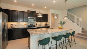 A modern kitchen with dark cabinets, stainless steel appliances, a granite countertop, and a central island with bar stools. - Elegant 5-bedroom Villa w Private Pool Near Disney (Davenport)