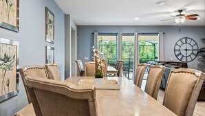 A dining area with a long table and chairs, a ceiling fan, and a large window with curtains. - Ultimate 8BR Family Fun in Resort Near Disney! (Kissimmee)