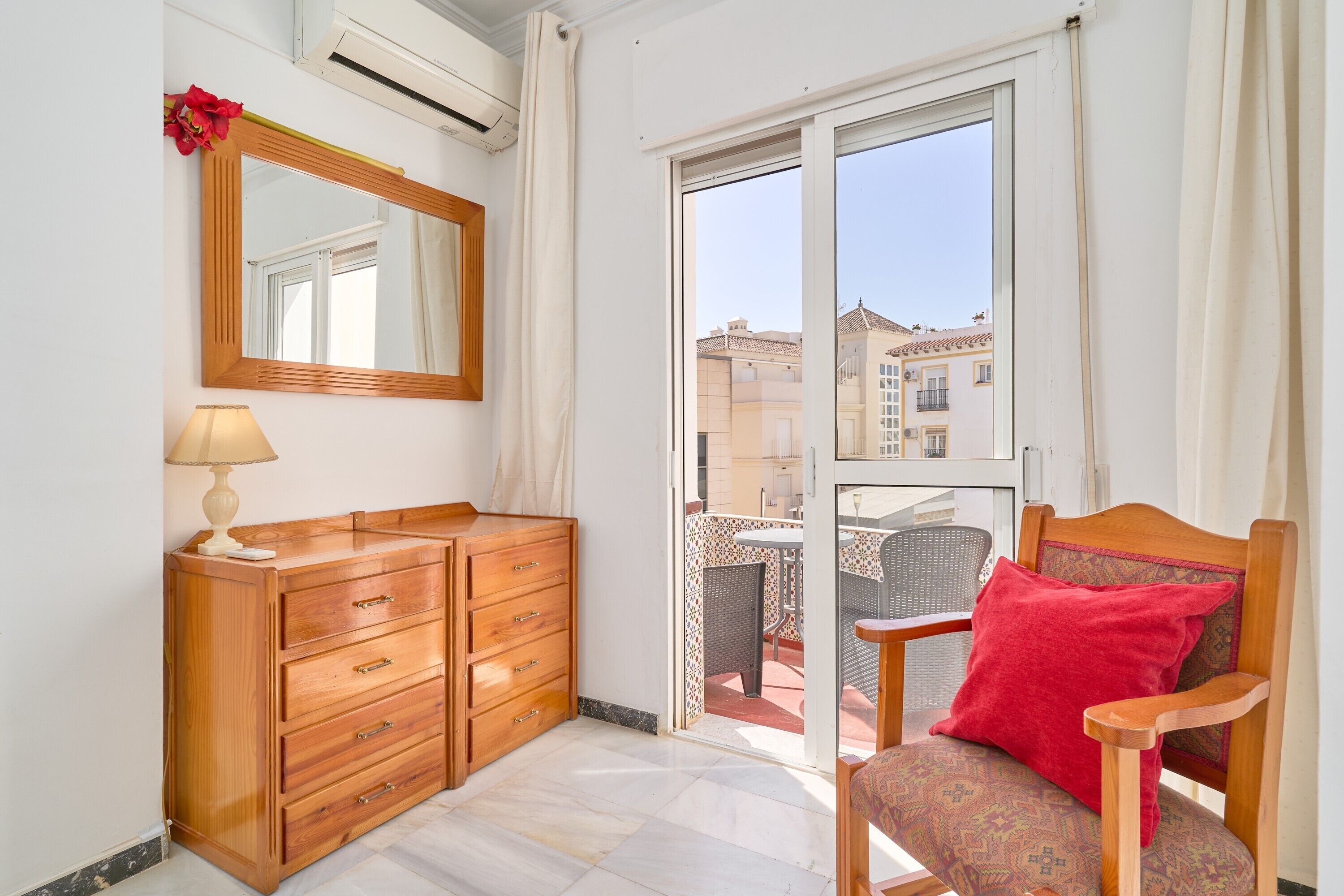 Una habitación con un aparador de madera, una silla con un cojín rojo, un espejo y vistas a edificios a través de una puerta de cristal.