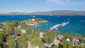Zona costera con puerto deportivo, barcos y playa con árboles y vehículos estacionados.