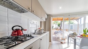 Una cocina moderna con armarios blancos, una estufa a gas y un hervidor rojo. Hay una zona de comedor con mesa y sillas, y una vista a un patio trasero con terraza de madera y valla blanca.