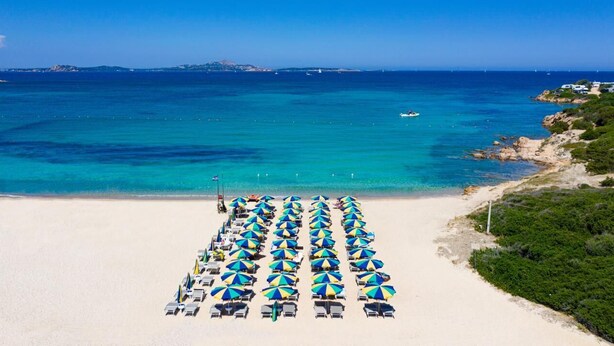 Una playa con filas de tumbonas y sombrillas, un mar azul cristalino y formaciones terrestres a la distancia.