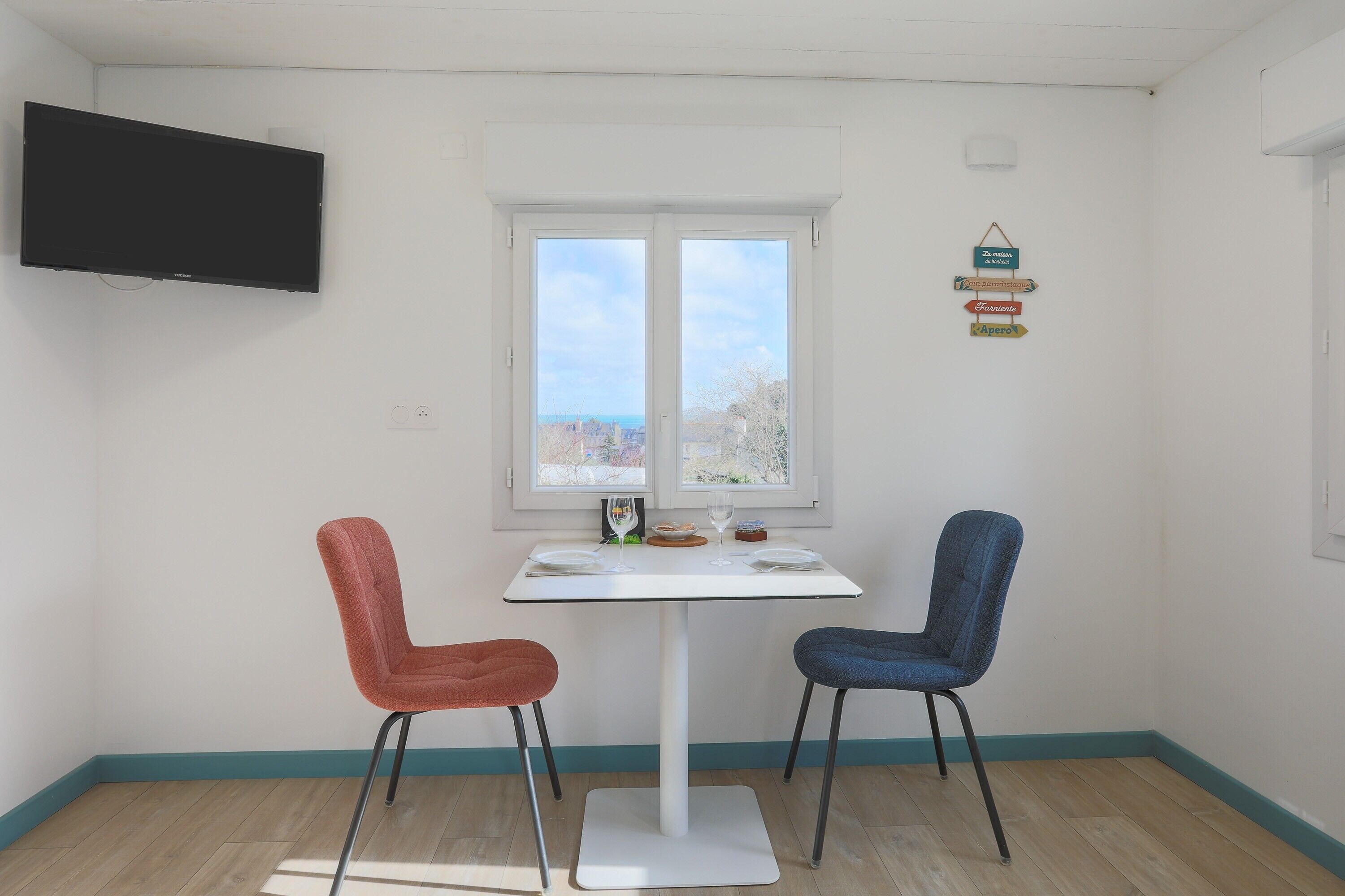 Quarto com mesa branca, duas cadeiras, televisão e janela com vista.